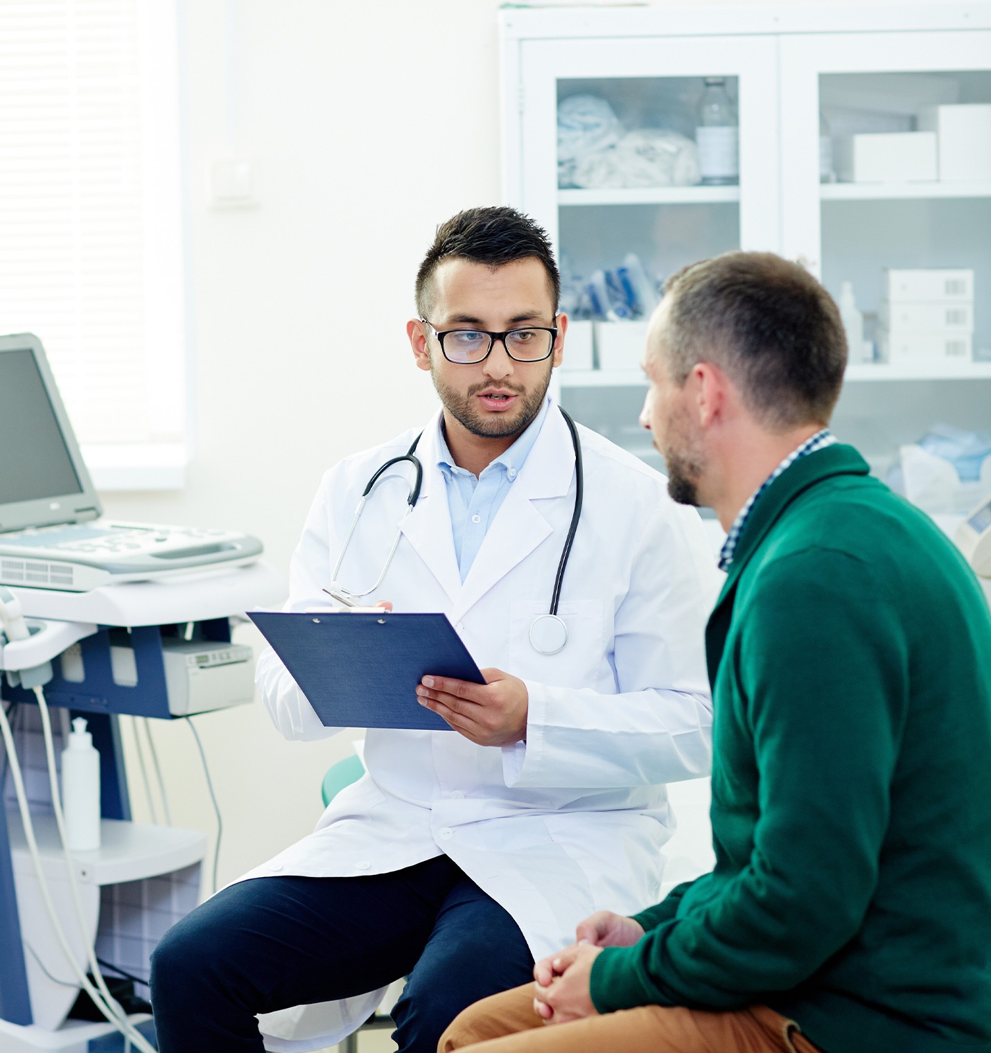 Ein Mann mit einem Kittel und Stethoskop um den Hals sitzt mit einem Patienten im Untersuchungsraum. Dabei hält er ein Klemmbrett in der Hand.