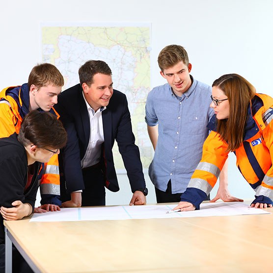 Fünf Personen beraten sich über einem Tisch auf dem mehrere Blätter liegen. Zwei der Personen tragen orange leuchtende Arbeitskleidung. Im Hintergrund hängt eine Karte.