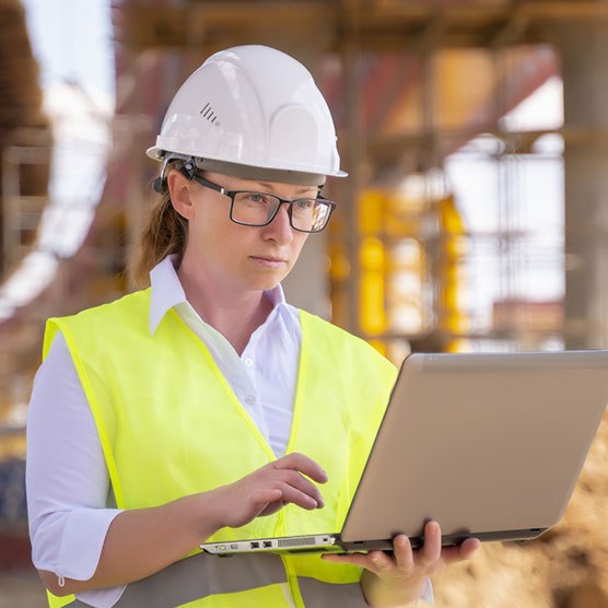 Eine Frau in Arbeitsschutzausrüstung steht mit ihrem aufgeklappten Laptop auf einer Baustelle.